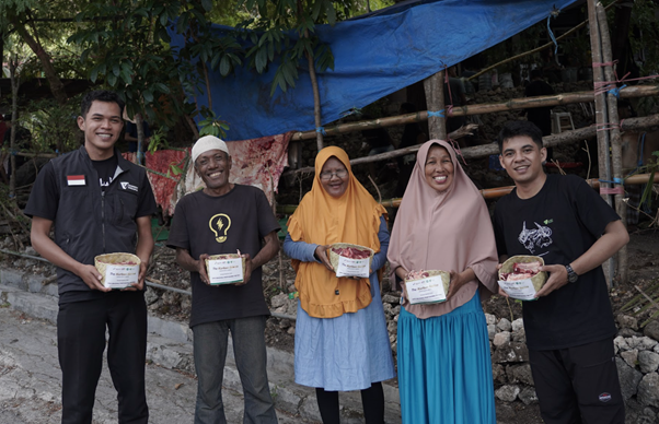 Five people stand in a row outdoors, each holding a packaged meal or bowl, in front of a blue tarp shelter.