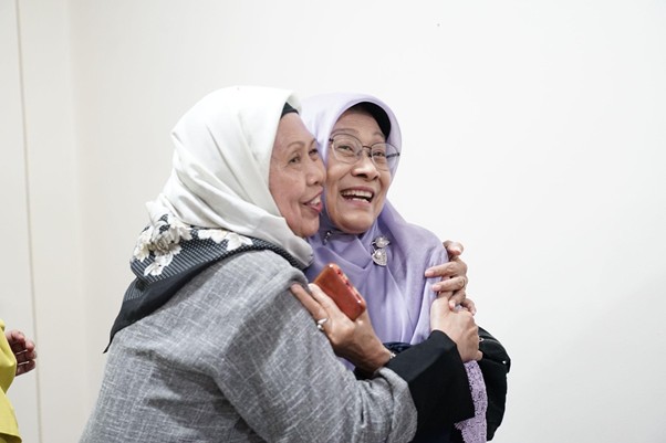Two elderly women hugging and smiling warmly, sharing a joyful moment in a bright room.