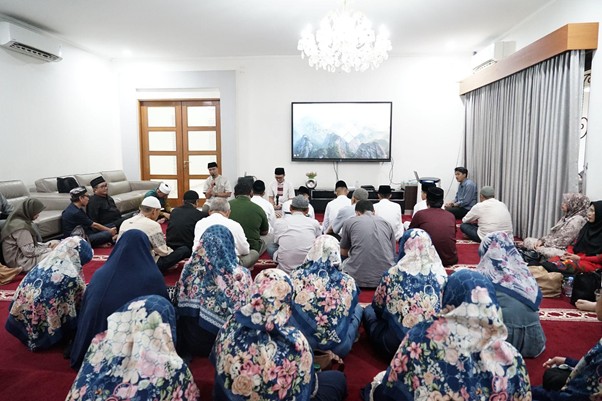 doa bersama keluarga iswadji 2 dompet dhuafa People seated on a red carpet in a bright living room for a community/religious gathering; women in floral headscarves sit in front, men behind, with a chandelier and mountain picture on the wall.
