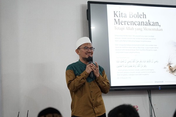 doa bersama keluarga iswadji 7 dompet dhuafa Speaker in a white cap and brown shirt holds a microphone beside a projected slide that reads 'Kita Boleh Merencanakan, Tetapi Allah yang Menentukan'.