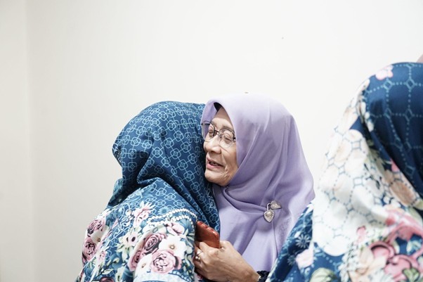 doa bersama keluarga iswadji 9 dompet dhuafa An elderly woman in a lavender hijab smiles as she hugs a person in a blue floral hijab; they stand indoors against a light background.