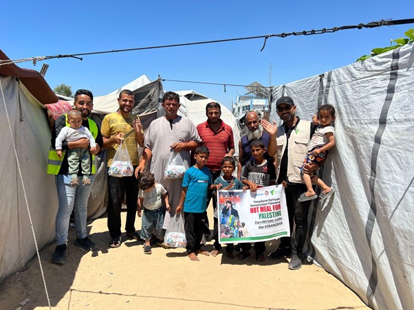 Group of volunteers and families pose together inside a temporary tent camp, holding bags of relief supplies and a banner supporting aid for Palestine.