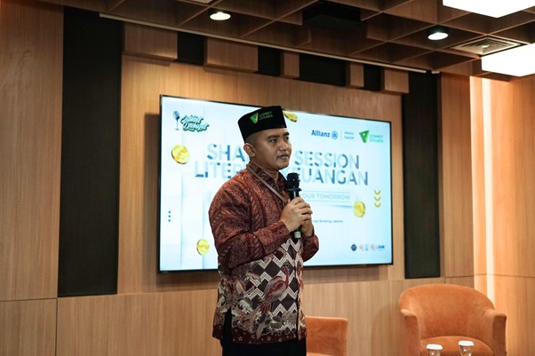 Man in a patterned batik shirt and black cap speaks into a microphone on a stage, with a presentation screen behind him showing event graphics.