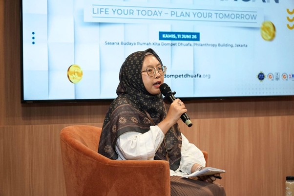 Woman in a patterned hijab and glasses speaks into a handheld microphone on a stage, holding papers, with a banner backdrop behind her.