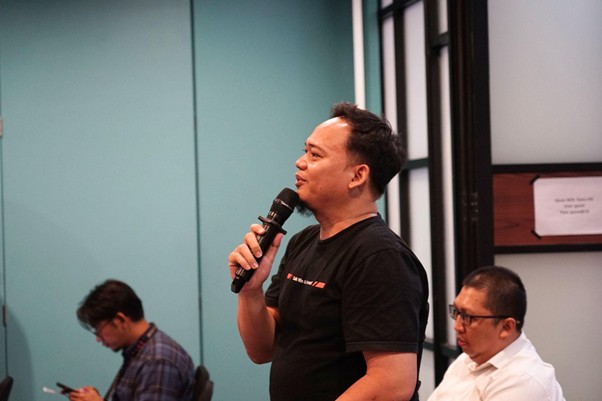 Man in a black t-shirt speaks into a handheld microphone at a meeting, teal wall behind him and two men seated in the background.