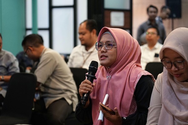 Woman in pink hijab and glasses speaks into a handheld microphone at a conference, holding a phone and notepad; attendees sit in the background.