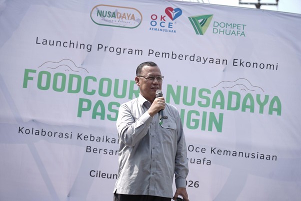 foodcourt nusadaya bogor 4 dompet dhuafa Man in a light gray shirt speaks into a handheld microphone on a stage, with a large banner and logos behind him.