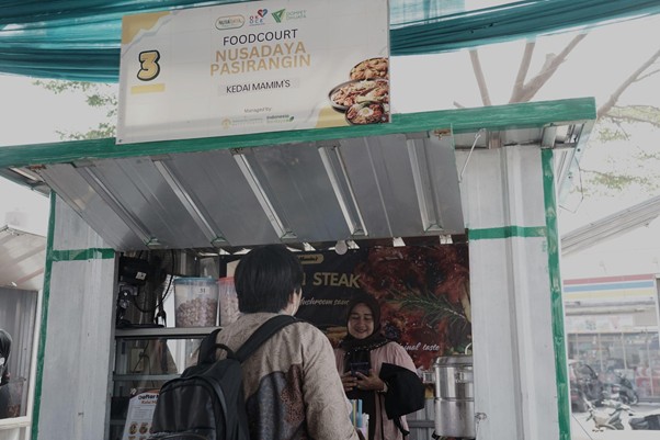 foodcourt nusadaya bogor 7 dompet dhuafa Customer with a backpack talks to a smiling female vendor at a food stall; a sign above reads 'Foodcourt Nusadaya Pasiringin' and shows stall number 3.