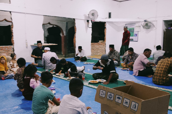 Large group seated on a blue plastic tarp inside a white-walled hall, eating a communal meal together on the floor.