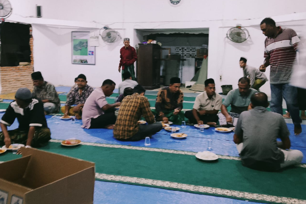 Group of men sitting on a blue mat on the floor, eating meals from plates and cups in a large hall.