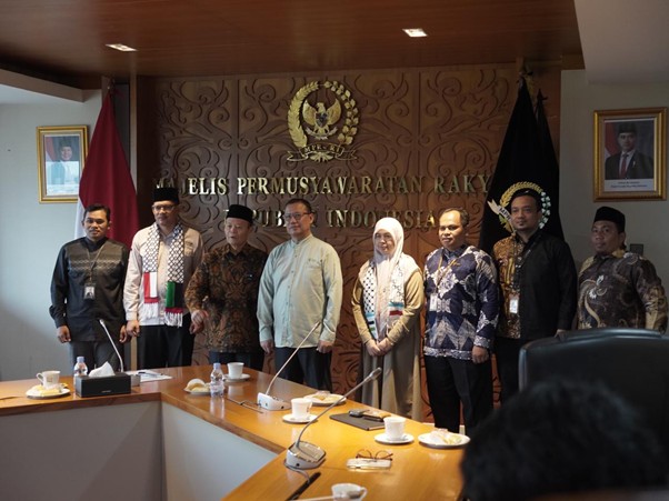 gpcikempr1 dompet dhuafa Ten people stand side by side in a formal meeting room, in front of a carved wooden wall with the Indonesian Garuda emblem and text, flanked by flags and portraits on the walls, all wearing name badges and traditional or business attire.