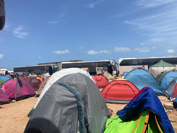 Colorful tents pitched on sandy ground with large buses in the background under a blue sky.