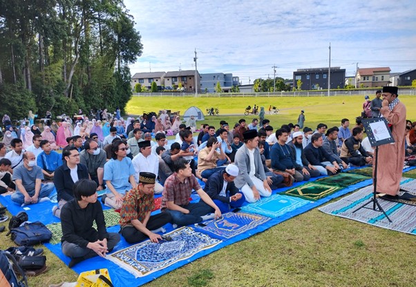 Large crowd seated on blue prayer mats on a sunny grassy field, listening to a speaker in traditional clothing at an outdoor religious gathering.