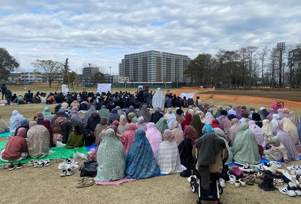 iduladha jepang 2 dompet dhuafa Large group of people wearing colorful robes sit on prayer mats outdoors, with shoes off nearby, on a grassy field against a stadium backdrop.