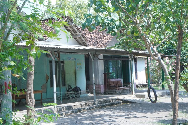 iduladha di rumah bu zahroh 4 dompet dhuafa A small, shaded porch area of a rustic building with a tiled roof and blue-green walls, surrounded by trees and a tire swing hanging from a tree branch