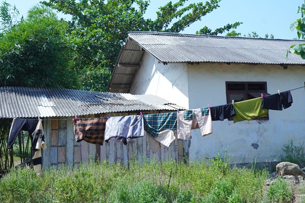 iduladha di rumah bu zahroh 6 dompet dhuafa White rural house with a corrugated metal roof; clothesline across the yard with colorful clothes drying, tall grass in front.