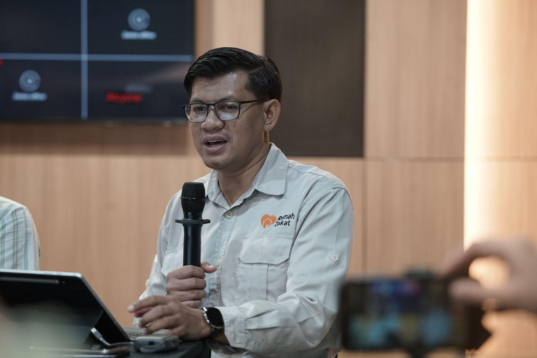 Man in a light-colored shirt with a logo on the chest speaks into a handheld microphone at a panel or conference room.