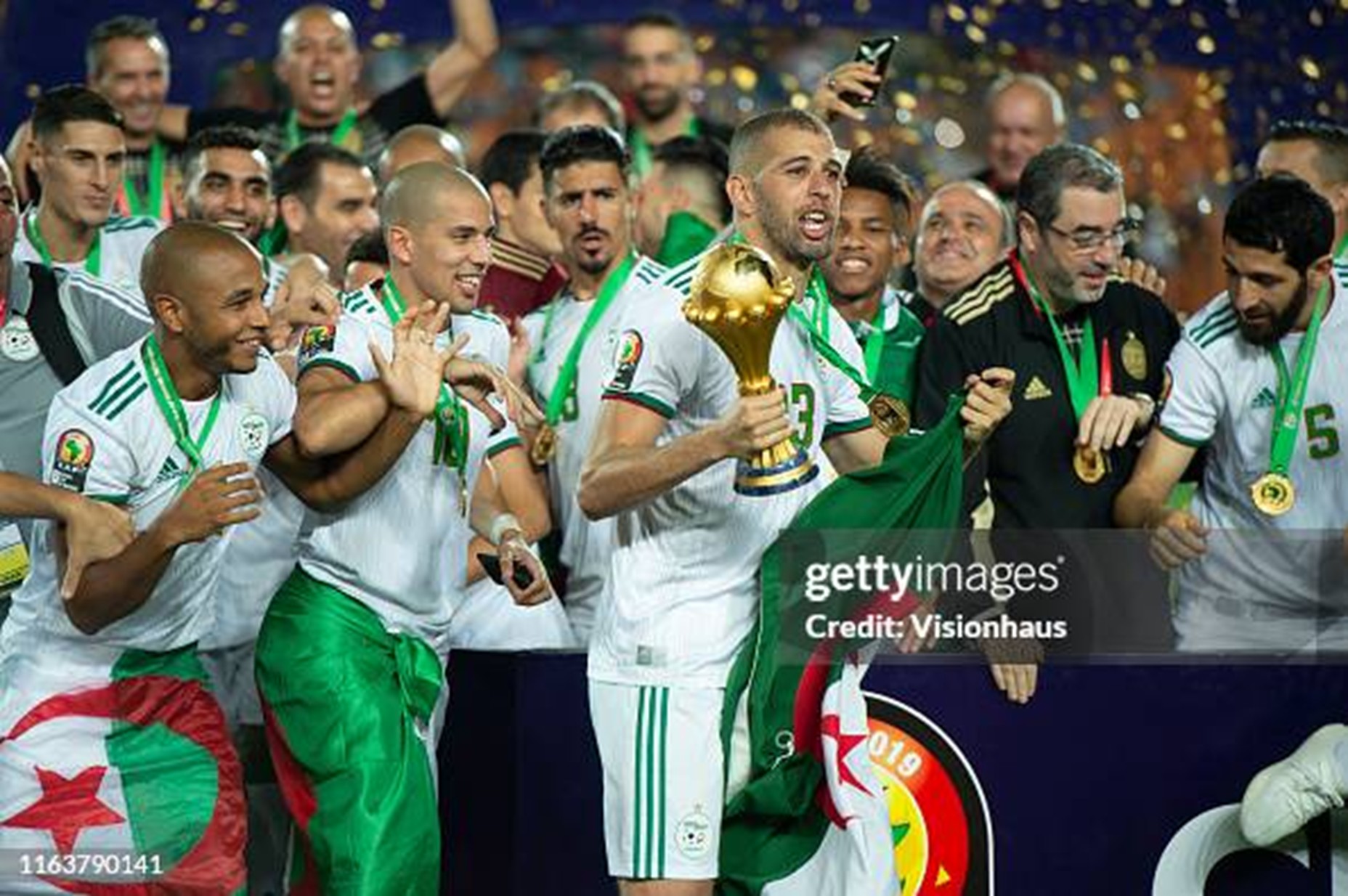 islam slimani 2 dompet dhuafa Algerian soccer players celebrate with a golden trophy, wearing white uniforms and green sashes, as confetti falls and fans cheer.