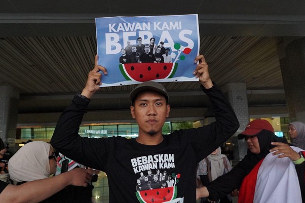 kedatangandelegasigsf204 dompet dhuafa Man in a black shirt raises a blue poster reading 'Kawan Kami Berbas' with a watermelon graphic, in a crowded indoor setting.