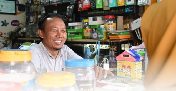 Smiling shopkeeper behind a cluttered counter with jars and merchandise in a small shop.