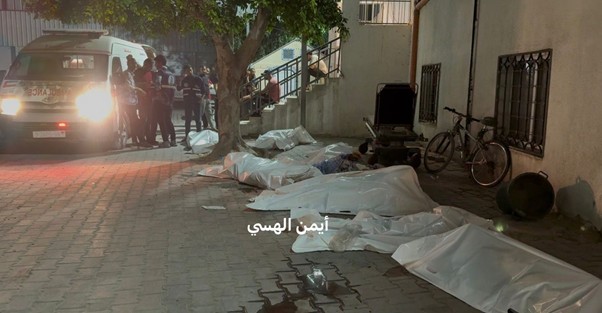 Night-time street scene with multiple white body bags lined along the pavement, emergency responders and an ambulance in the background.