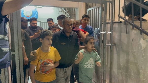 Man leading two children through a metal gate as a crowd of people watches in a narrow hallway; Arabic text appears on the right side.