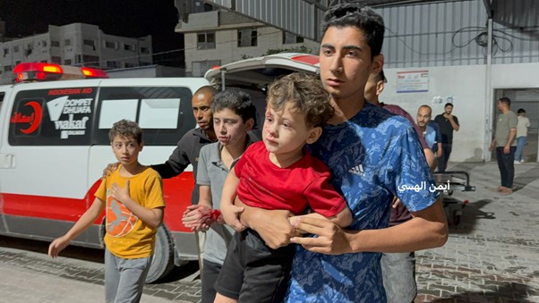 Young man in a blue shirt carries a small boy in a red shirt, with bystanders and an ambulance in the background at night.