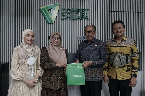 Four people stand indoors in front of a Dompet Dhuafa sign; a man and a woman exchange a green donation bag.