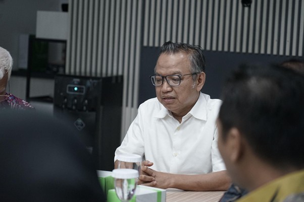 Middle-aged man in a white shirt and glasses sits at a conference table during a meeting, hands clasped, focused on discussion.