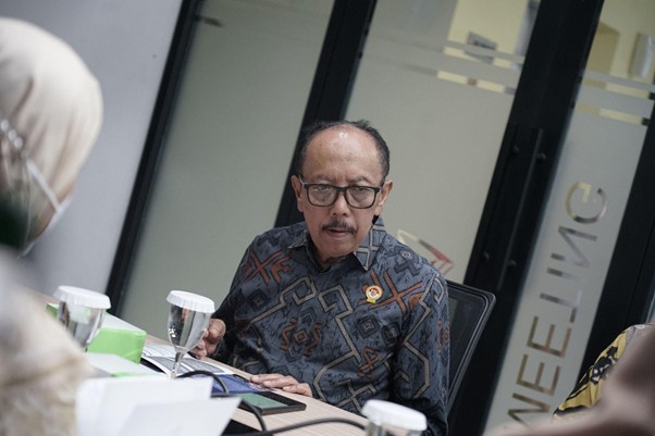 Man with glasses in a batik shirt sits at a meeting table, looking at a tablet with cups and a notepad nearby.