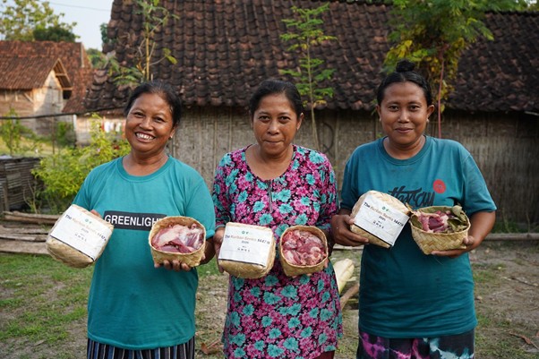 Three women stand outdoors, each holding a woven basket with meat portions, smiling at the camera.