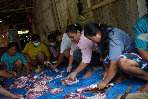 Community group processing raw meat on a blue tarp inside a wooden shed, sorting and cutting meat pieces together.