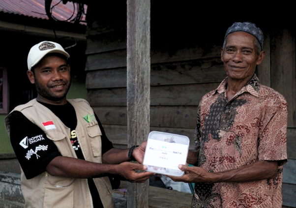 kurban hmns di maluku 1 dompet dhuafa Two men exchange a white wrapped package outside a wooden house; one wears a beige vest and cap, the other a patterned shirt, both smiling.
