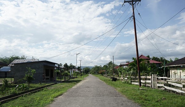 kurban hmns di maluku 2 dompet dhuafa Rural dirt road in a small village, flanked by modest houses, fences, and overhead power lines under a partly cloudy sky.
