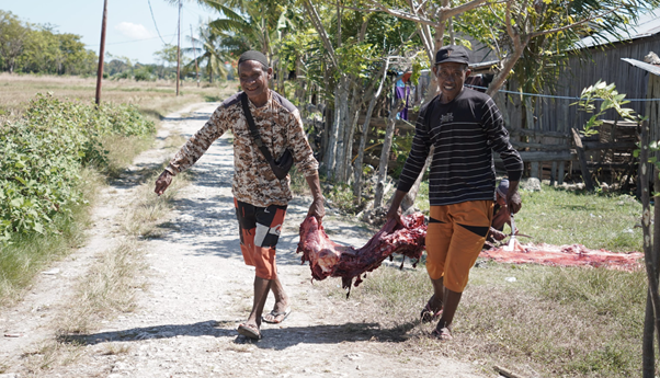Two men walk down a rural dirt road, carrying a large butchered animal carcass between them.