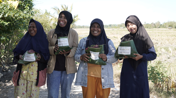 Four smiling women wearing hijabs stand outdoors, each holding green-wrapped food parcels or boxes in a sunny rural setting.