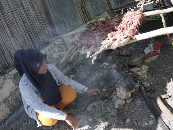 Woman in a blue headscarf crouches beside a small fire, cooking kebabs skewered on sticks over smoky flames near a corrugated metal wall.