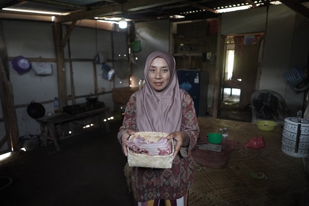 Woman in a hijab holding a wrapped loaf in a small, rustic home kitchen interior.