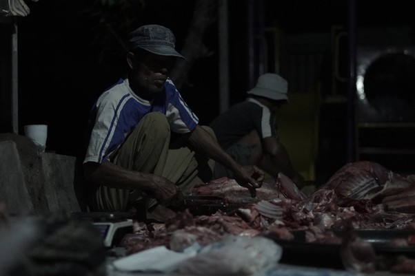 Man in a cap slicing raw meat at a dimly lit market stall, with another worker in the background.