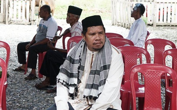 Central man in traditional attire (white robe, black cap, scarf) sits among others on red plastic chairs at an outdoor gathering; gravel ground and fence in background.