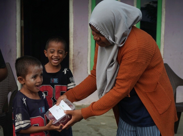 Woman in a hijab and orange cardigan hands a gift box to a smiling boy, as another child laughs nearby.