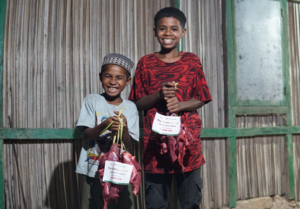 Two smiling boys stand outside a wooden house, each holding a bag of fresh vegetables.