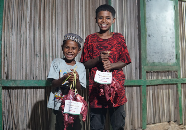 kurban rois ojk 1 dompet dhuafa Two smiling boys stand outside a wooden house, each holding a bag of fresh vegetables.