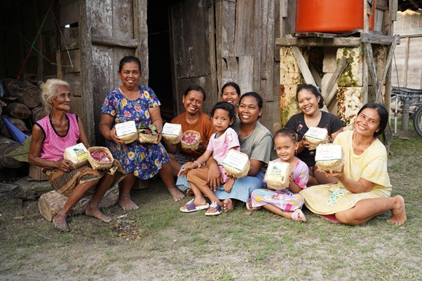 kurbanroisojk11 dompet dhuafa Smiling women and children sit on the ground outside a weathered wooden house, each holding a carton of eggs.