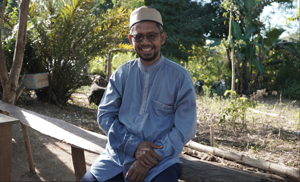 kurban rois ojk 12 dompet dhuafa Man wearing a light blue traditional tunic and kufi cap sits on a wooden bench outdoors, smiling at the camera in a sunny garden setting.