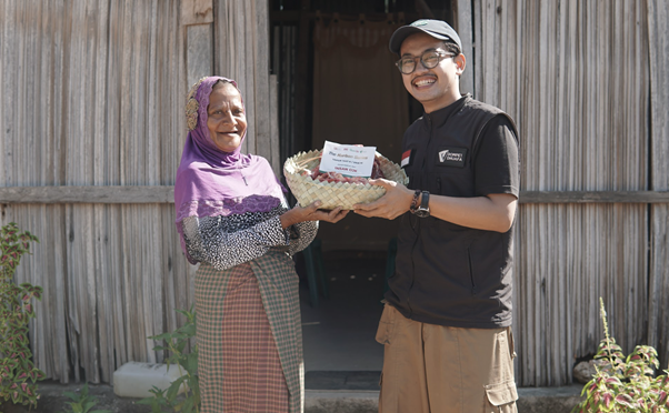 kurban rois ojk 17 dompet dhuafa Volunteer hands a gift basket to an elderly woman standing outside a wooden home; both smile at the camera.