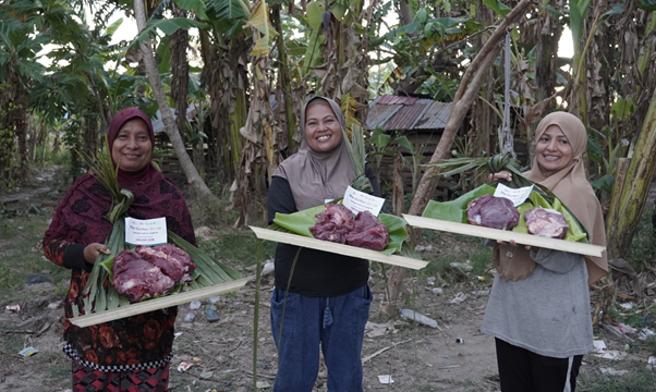 kurban rois ojk 18 dompet dhuafa Three women wearing hijabs stand outdoors, each holding a wooden tray with portions of raw meat on banana leaves.