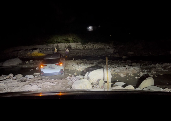 kurban rois ojk 2 dompet dhuafa White SUV stranded on a rocky riverbank at night, with two people nearby and headlights on; water and large boulders surround the scene.