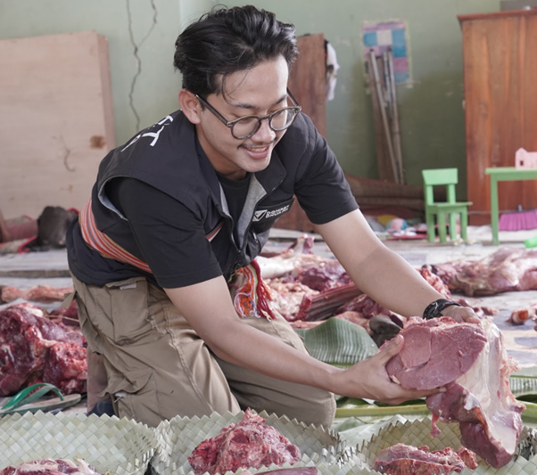 kurban rois ojk 5 dompet dhuafa Man with glasses kneels in a meat market, handling a large raw cut of meat among other carcasses and bones in a cluttered workspace.
