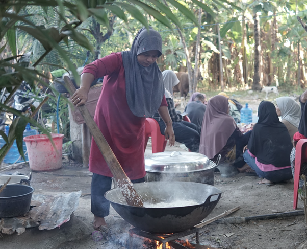 kurban rois ojk 7 dompet dhuafa Woman wearing a gray headscarf stirs a large wok over an open fire outdoors, with people seated nearby under trees in the background.